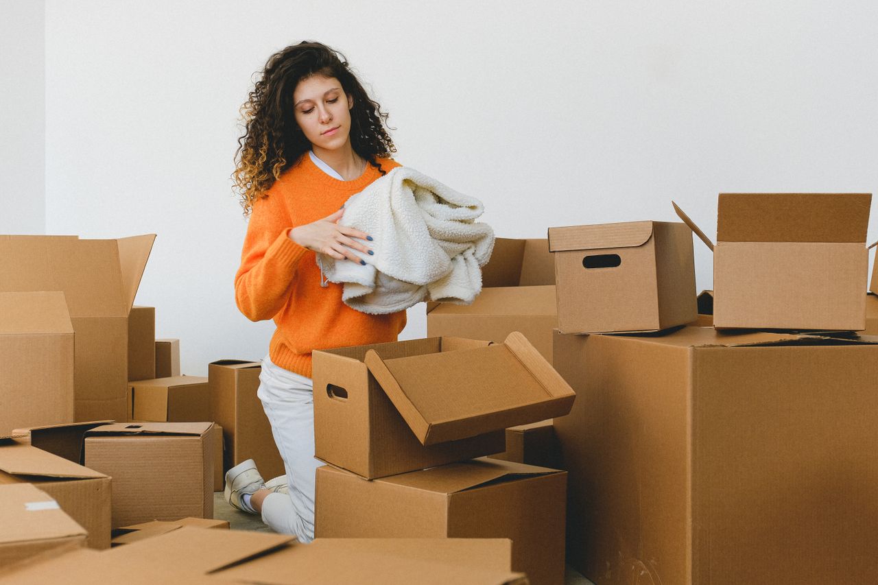 expertise-01 Jeune femme concentrée aux cheveux bouclés, agenouillée au sol et rangeant des vêtements dans des cartons avant de déménager dans un nouvel appartement